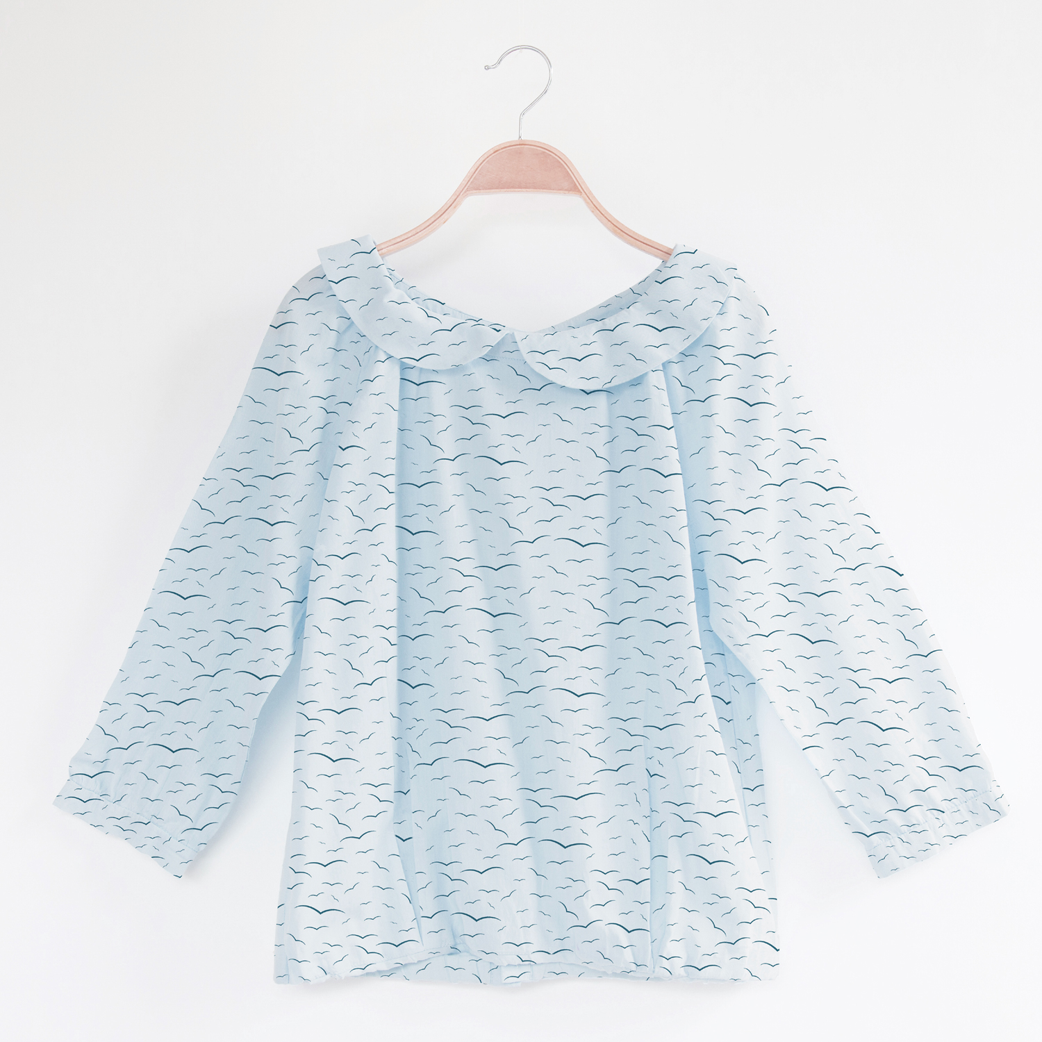 Image of a pretty linen blouse hanging on a wooden hanger. The blouse features "Gulls", a pattern from the Quayside collection.