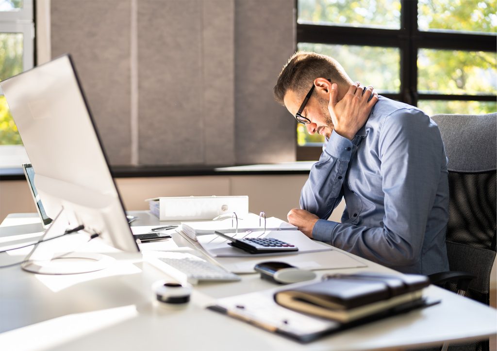 An image of a man sitting at his desk with one hand cradling his neck as though he is experiencing some discomfort. Office Wellness.