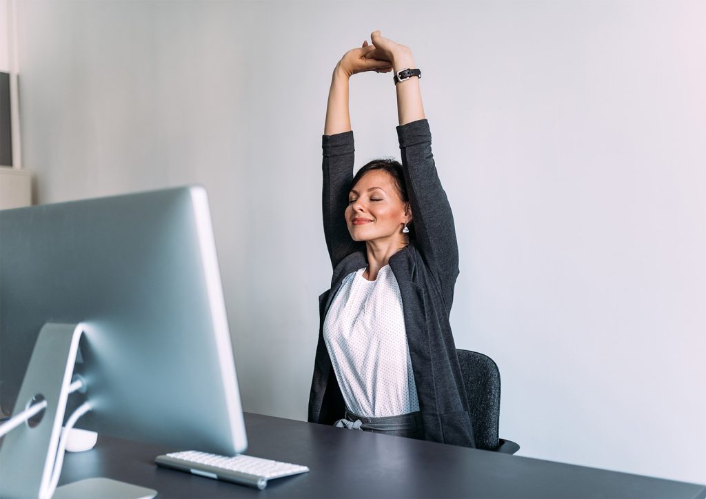 An image of a woman sitting at her desk stretching both her arms above her head and taking in a deep breath.