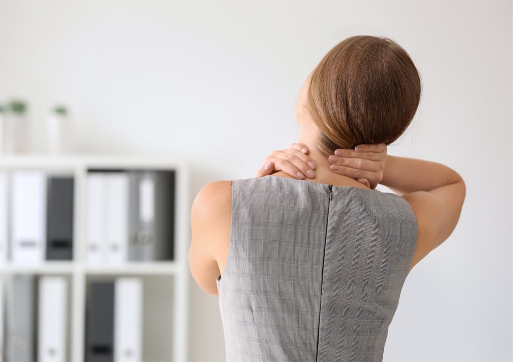 Image of a woman in an office setting with her back (us) the camera with her hands reaching around to the back of her head, appearing to be in discomfort. Office Wellness.